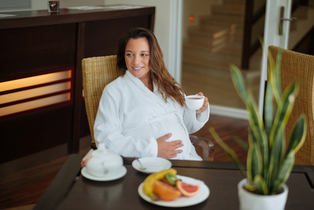 schwangere Frau im Bademantel sitzt an einem Tisch und genießt eine Tasse Tee